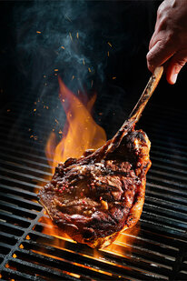 Eine Person wendet ein brutzelndes Steak auf einem brennenden Grill, wobei Rauch aufsteigt.