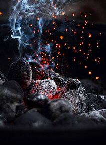 Glühende Holzkohleglut mit aufsteigendem Rauch und Funken in einer dunklen Umgebung.