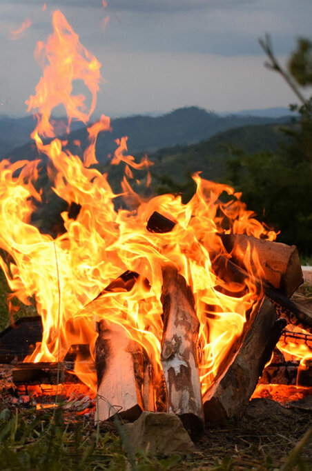 Lagerfeuer mit intensiven Flammen vor einem abendlichen Himmel, mit Silhouetten von Menschen, die sich in einer natürlichen Umgebung darum versammelt haben.