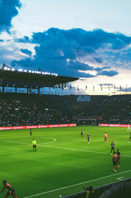 Ein Fußballspiel in einem Stadion mit über das ganze Feld verstreuten Spielern, leuchtend grünem Gras und einer Menschenmenge unter einem bewölkten blauen Himmel.