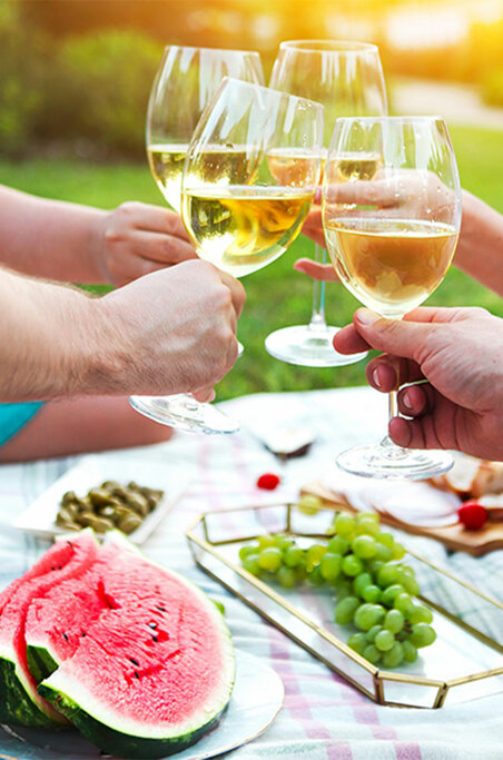 Zwei Personen essen im Freien. Ihre Hände greifen sichtbar nach dem Essen; sonnenbeschienener Tisch mit Sommerfrüchten, Brot und Wein.