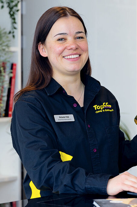 Eine Frau mit Pferdeschwanz, eine schwarze Uniform mit gelben Logos tragend, lächelt und steht in einer Industrieumgebung.