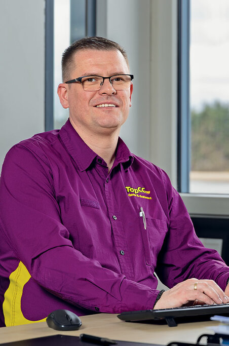 Ein Mann in lila Hemd und Brille sitzt mit einem Laptop an einem Schreibtisch und lächelt in die Kamera, während durch die Fenster hinter ihm Licht hereinströmt.