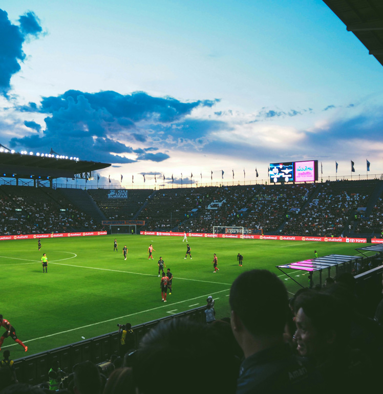 Menschenmenge schaut sich bei klarem Abendhimmel ein Fußballspiel in einem Stadion an.