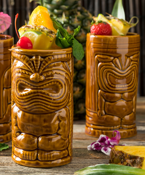Zwei Tiki-Becher gefüllt mit tropischen Cocktails, garniert mit Früchten und Blumen, auf einem Holztisch.