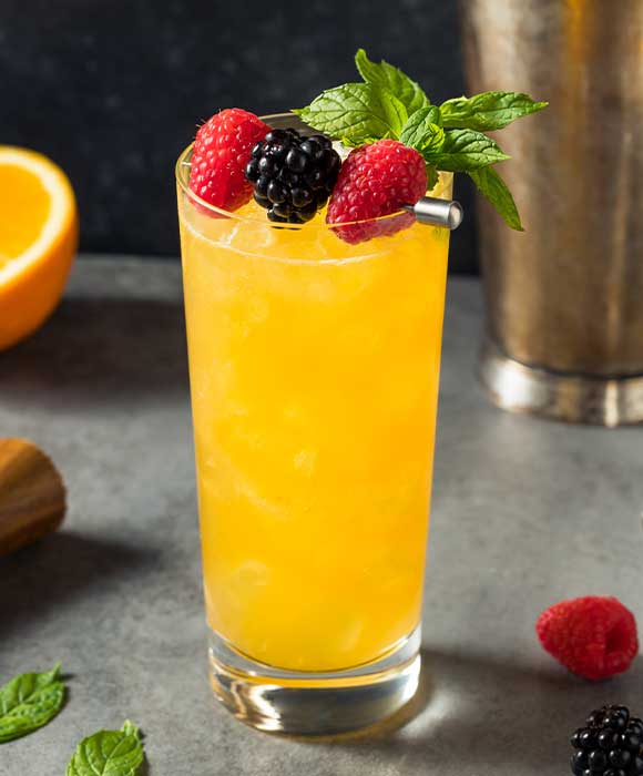 Ein gekühltes Glas Orangensaft, garniert mit frischen Beeren und Minze, auf einem Tisch mit Zitrusfrüchten im Hintergrund.