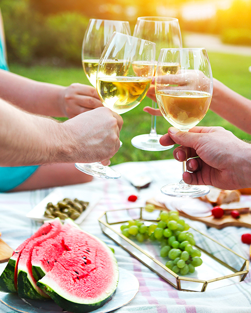 Nahaufnahme von Händen, die an einem sonnigen Tag mit Weingläsern über einem mit verschiedenen Speisen gedeckten Picknicktisch anstoßen.