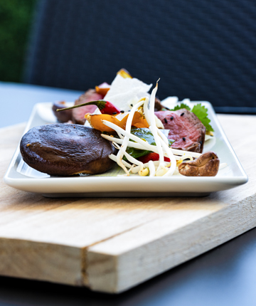 Ein Gourmetgericht bestehend aus einer Scheibe Fleisch mit Beilagen auf einem weißen Teller, angerichtet auf einem Holztisch.