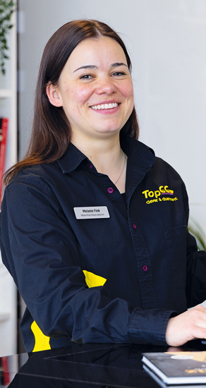 Eine lächelnde Frau mit langen Haaren, eine schwarze Uniform mit gelben Details tragend, hält in einem Innenbereich ein Tablet in der Hand.